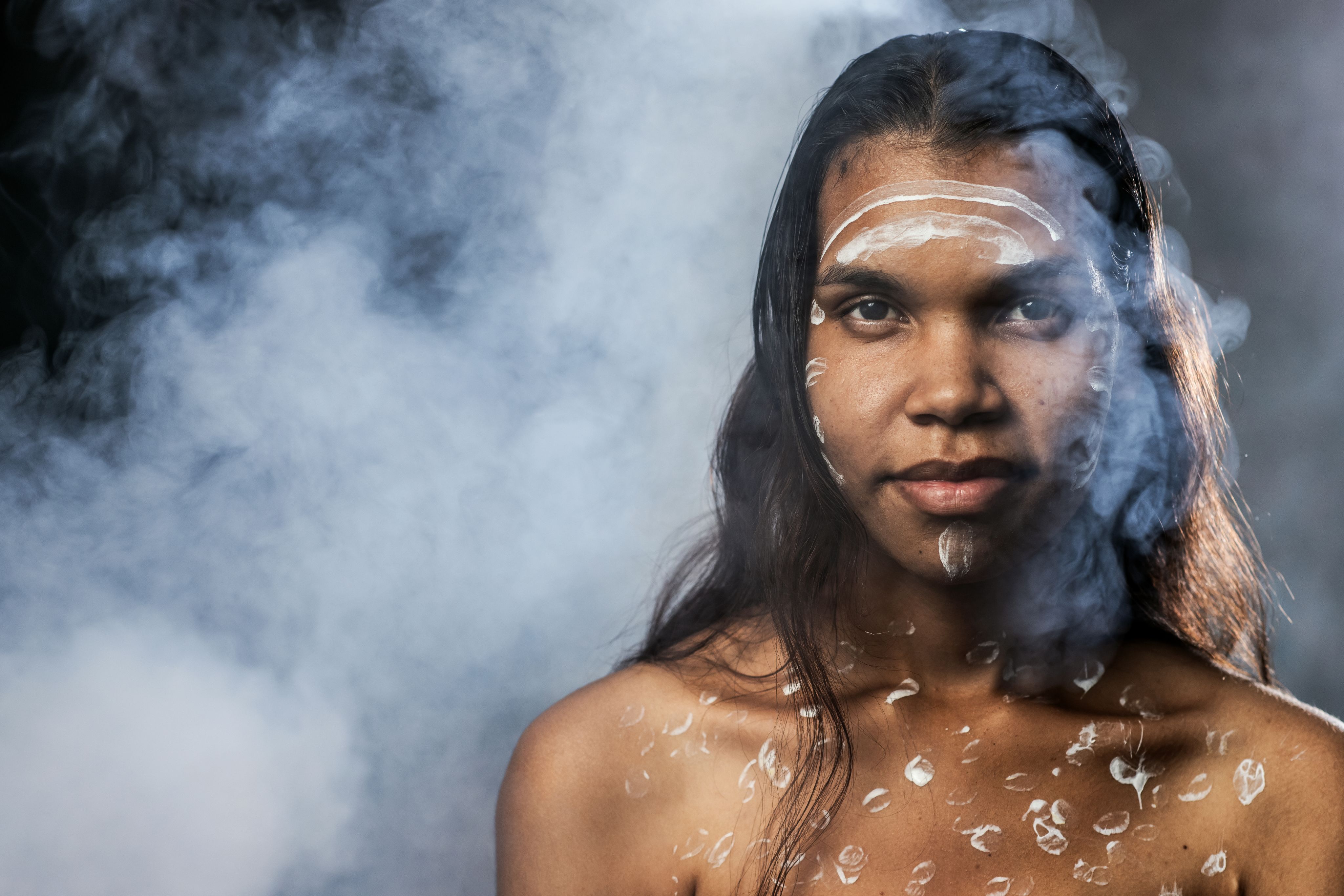 A woman is surrounded by smoke and has body paint on her face