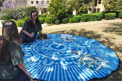 Two figures sitting on the edge of a blue, patterned, circular mat.