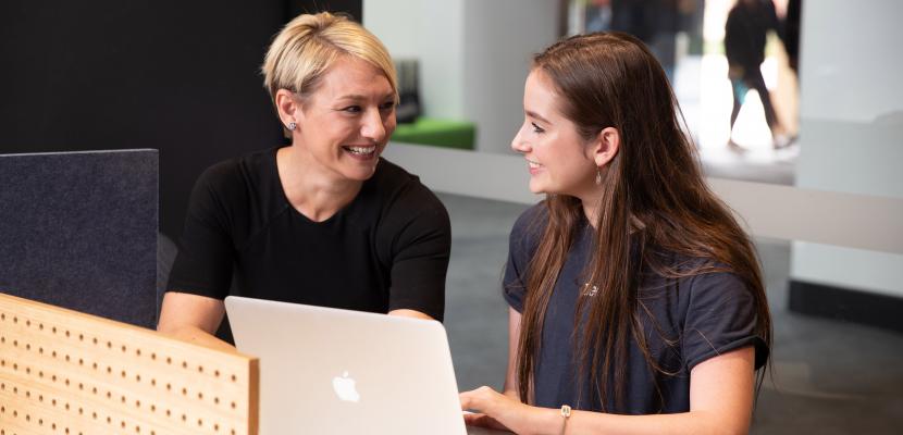 A female student and female teacher sitting town smiling in conversation with a Macbook laptop open on a desk in front of them.