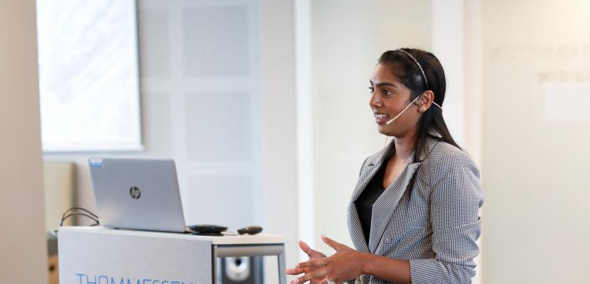 Meera Klemola stands in front of a lectern and a laptop, presenting