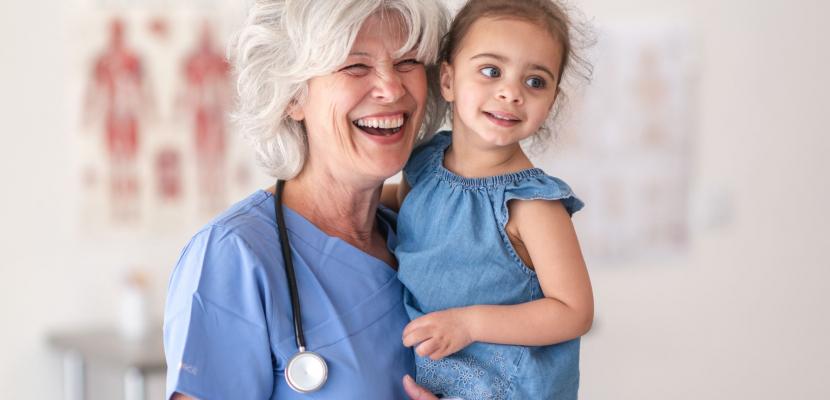 A doctor wearing a stethoscope holds a child and smiles at the camera