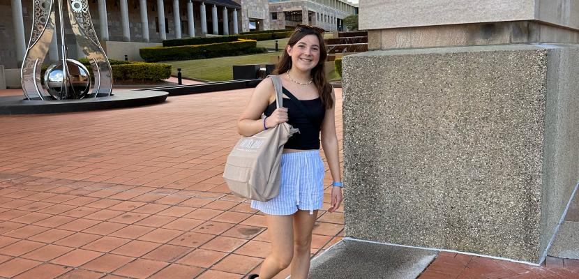 A girl with brown hair is smiling under the Bond University Arch with the Limitless sculpture in the background