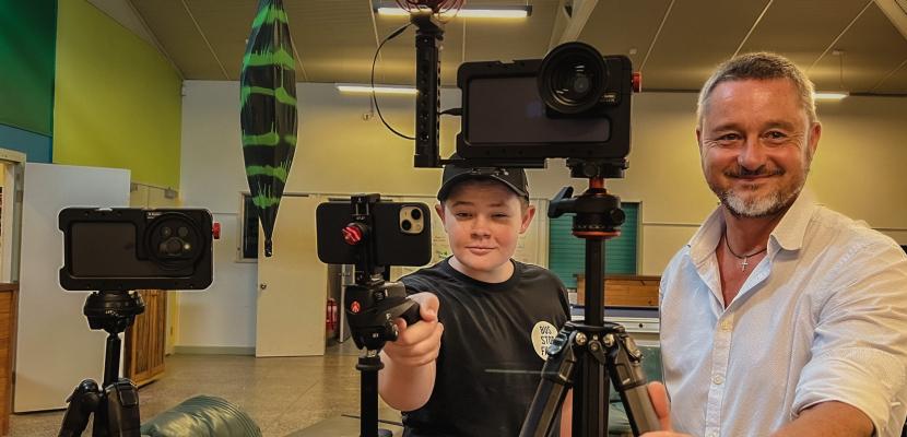 Rob and one of the young crew members stand behind iPhones on tripods, smiling.