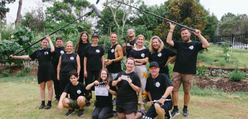 A film crew of about 15 people stands outside holding mics and smiling in black t-shirts