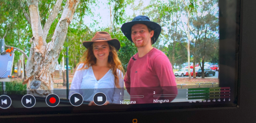 An image of a camera monitor. On the screen are two people wearing Akubra hats, smiling, with trees in the background.