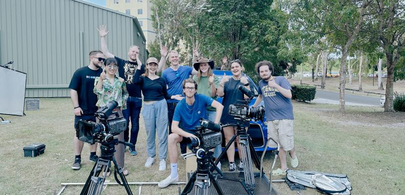 A group of students stand outside in front of trees and a shed, smiling. There is camera equipment in the foreground.