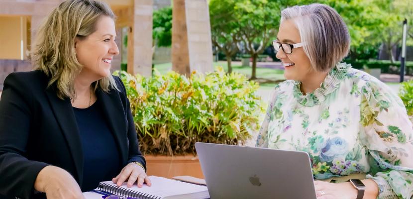 Natalie Hick and Michelle Peden work at an outside table. They are looking at each other and smiling.