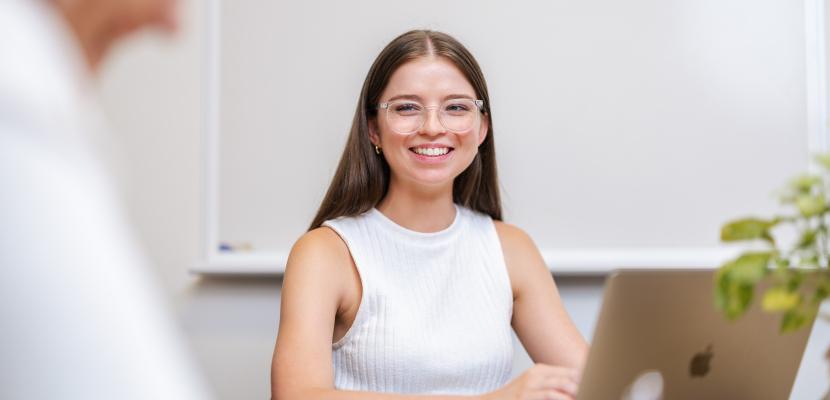 A young woman is sitting behind a laptop, smiling. 