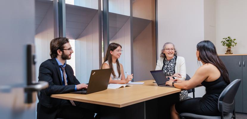 Four people in business attire with laptops and notepads sitting around a office desk