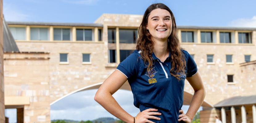 A young woman is standing in front o f a sandstone building with her hands on ehr hips and smiling.