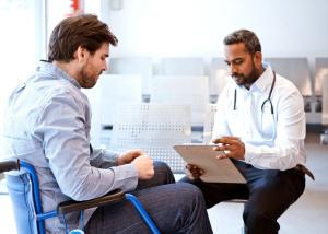 Doctor holds a clipboard speaking with a patient in a wheelchair