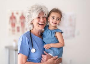 A doctor wearing a stethoscope holds a child and smiles at the camera