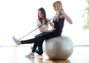 One women sits on an exercise ball using a resistance band to hold her leg up, another woman kneels beside her supporting her by holding her waist