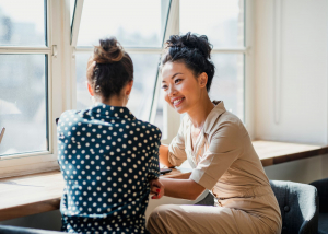 Two women sit at a table, one is facing away from the camera and one is facing towards the camera smiling at the other woman