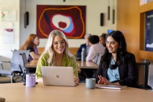 Two women sit at a desk in front of a laptop